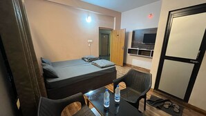 Une petite chambre comprenant un lit, deux chaises, une petite table avec des bouteilles d’eau, un téléviseur et une armoire.
