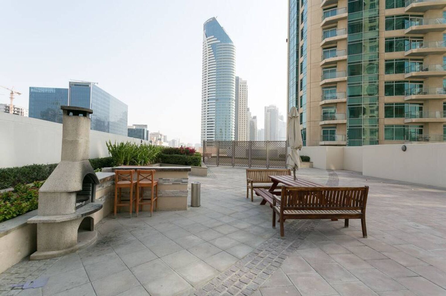 An outdoor patio area with a stone barbecue, wooden benches, and a view of modern high-rise buildings.