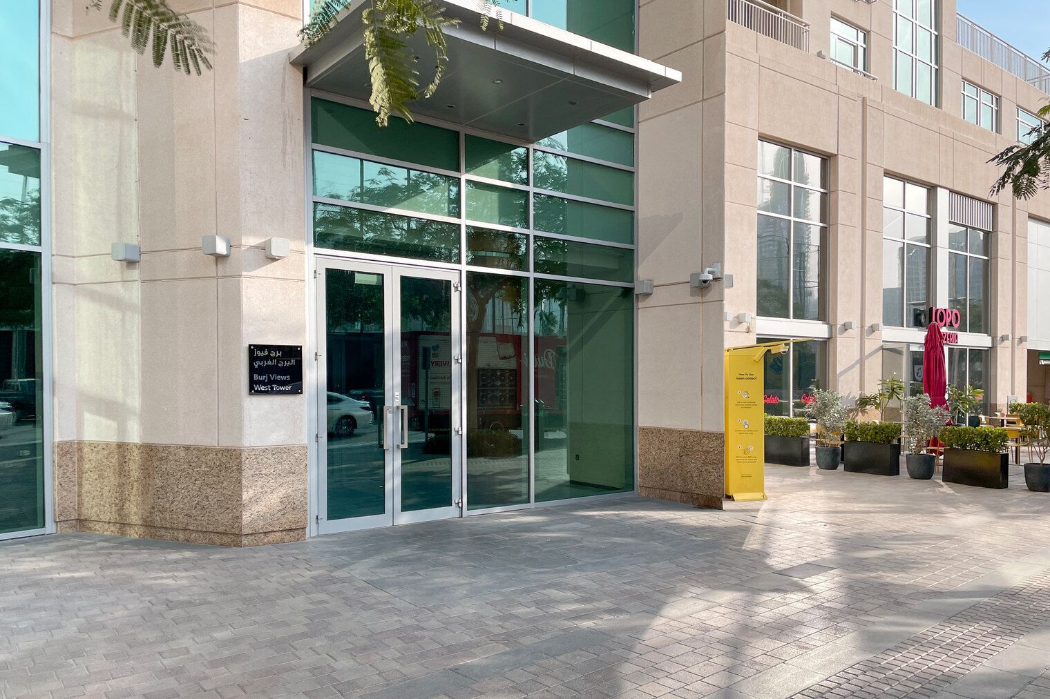 Ingresso di un edificio moderno con porte in vetro, un cartello con la scritta 'West Tower' e area esterna con posti a sedere e piante.