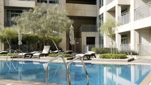 Une piscine entourée de chaises longues et de parasols, elle-même bordée d’un immeuble à plusieurs étages doté de balcons.