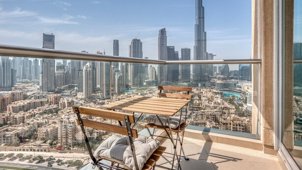 Un balcon avec une table et des chaises en bois, donnant sur un paysage urbain composé d’immeubles de grande hauteur et d’un gratte-ciel imposant.