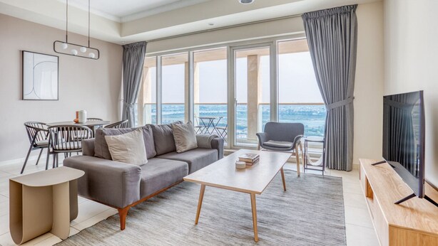 A modern living room with a sofa, coffee table, and TV, large windows with curtains, and a view of the city.