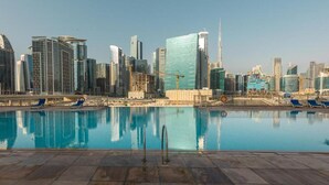 A cityscape with modern high-rise buildings, a clear sky, and a swimming pool with lounge chairs.