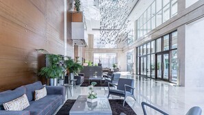 Un hall d’entrée spacieux au design moderne, avec un canapé gris, une table basse et plusieurs fauteuils. On y aperçoit également des plantes en pot et un escalier au fond.
