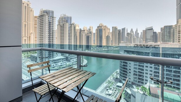 Un balcon avec un banc en bois et une vue sur un paysage urbain, avec de hauts immeubles et une étendue d’eau.