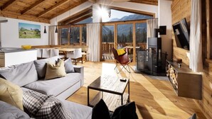 Een ruime woonkamer met een houten plafond, een bank, een salontafel en een televisie. Grote ramen bieden uitzicht op de omgeving.