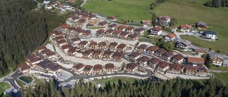 An aerial view of a residential area with houses clustered around a central road, surrounded by forested areas.