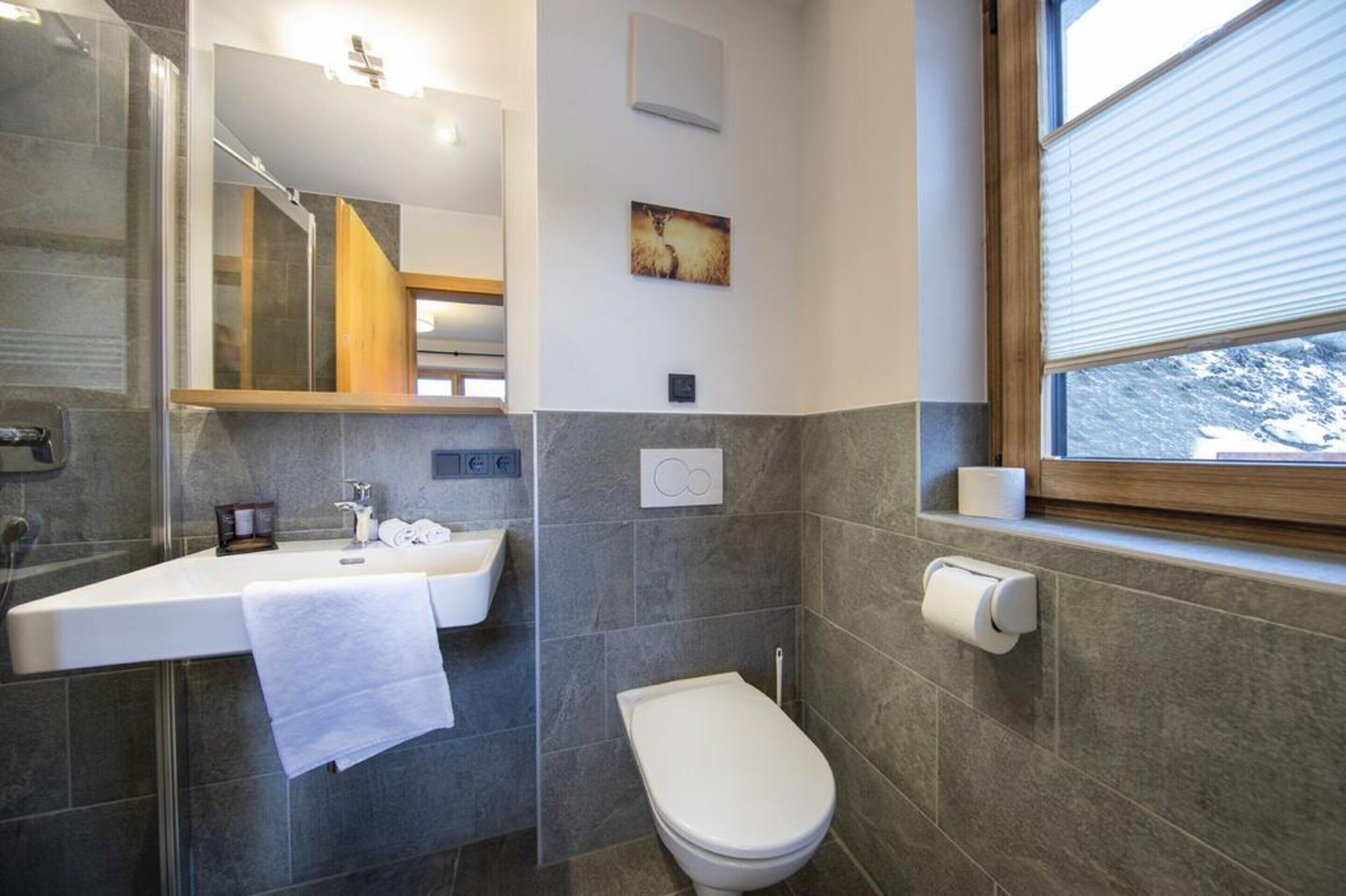 A modern bathroom with a glass shower enclosure, a white sink with a towel, a toilet, and a window with blinds.