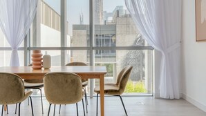 Ein moderner Essbereich mit Holztisch und -stühlen, großen Fenstern mit durchsichtigen Vorhängen und Blick auf Stadtgebäude.