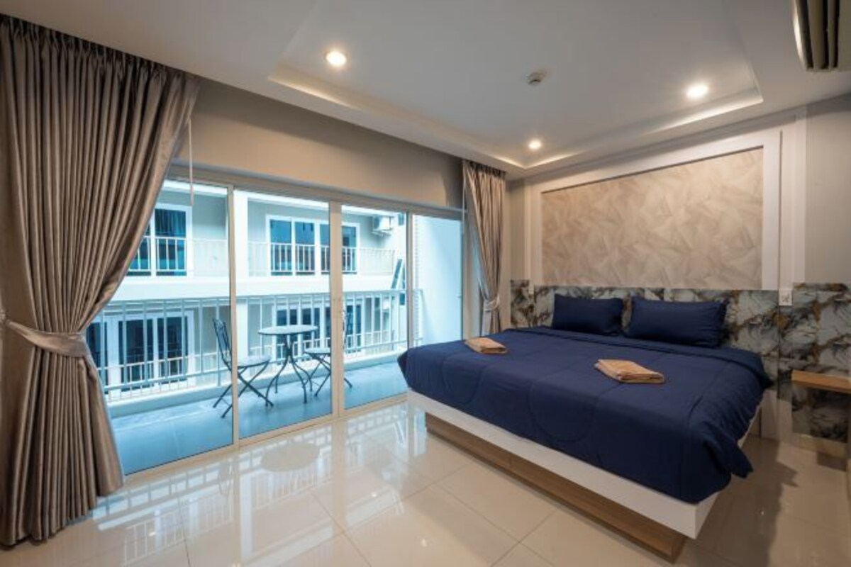 A modern bedroom with a large bed, a balcony with a table and chairs, and a view of a building.
