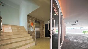 Um saguão de hotel com uma escadaria que leva ao primeiro andar, uma área de recepção e uma porta de vidro com um aviso de boas-vindas.