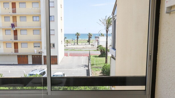 A room with a large window overlooking a beach and a building. There is a table with plates and glasses, and a bench with cushions. - St Cyprien beautiful cozy apartment sea view (Saint-Cyprien)