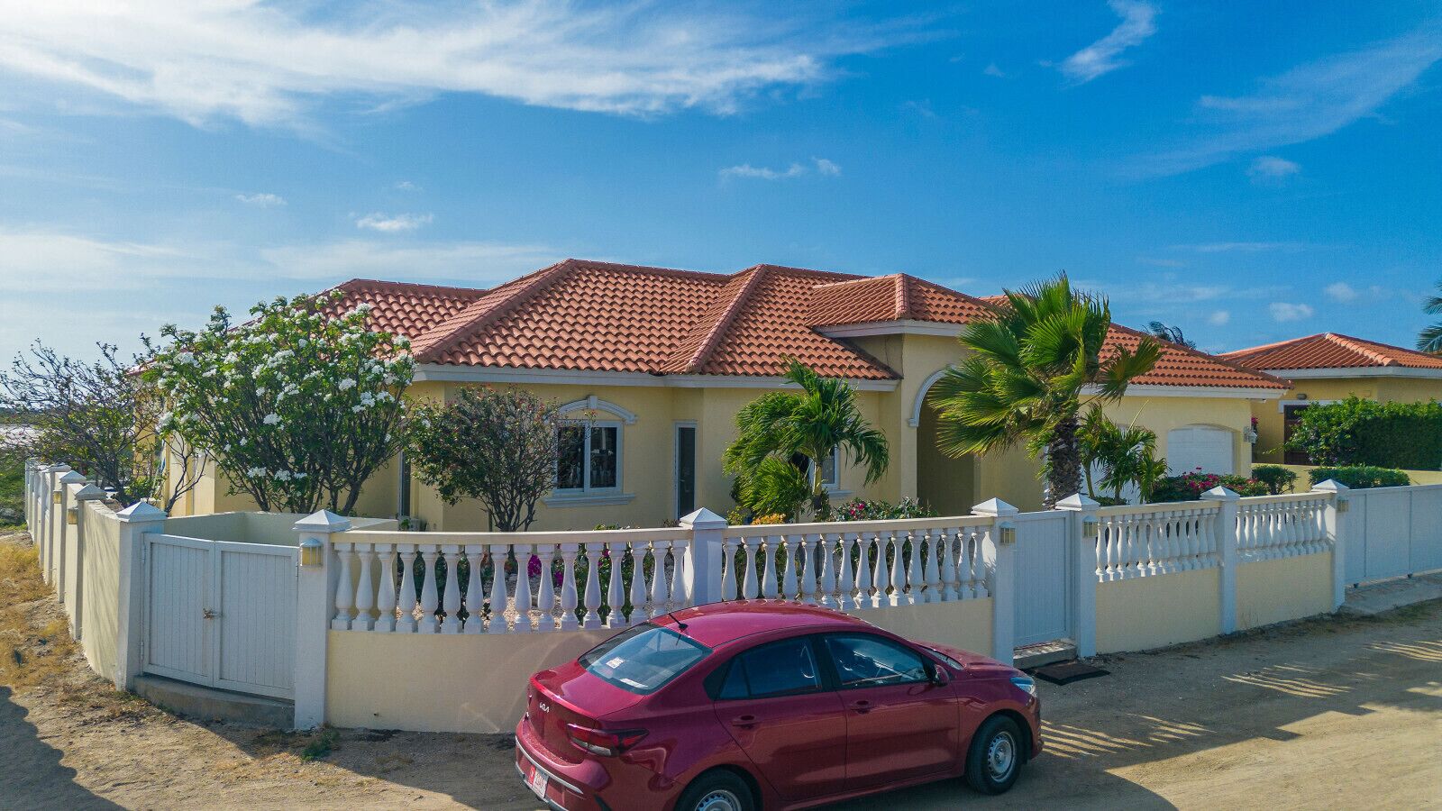 Uma casa de dois andares com telhado de telhas vermelhas, cerca branca de piquete e um carro vermelho estacionado na frente.