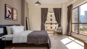 Ein modernes Hotelzimmer mit einem großen Bett, einem Sessel, einem Schreibtisch und Blick auf die Stadt durch große Fenster.