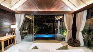 A modern bedroom with a glass door leading to a pool area, wooden ceiling, and a view of tropical plants.