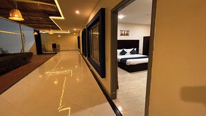 Un couloir mène à une chambre qui contient un lit, une table de chevet et un téléviseur fixé au mur. Le couloir est doté d’un sol carrelé et d’un plafond en bois avec des luminaires suspendus.