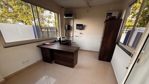 Une petite pièce bien éclairée, avec un bureau, un ordinateur et un téléviseur fixé au mur. Une fenêtre laisse entrer la lumière naturelle et offre une vue sur les arbres à l’extérieur.