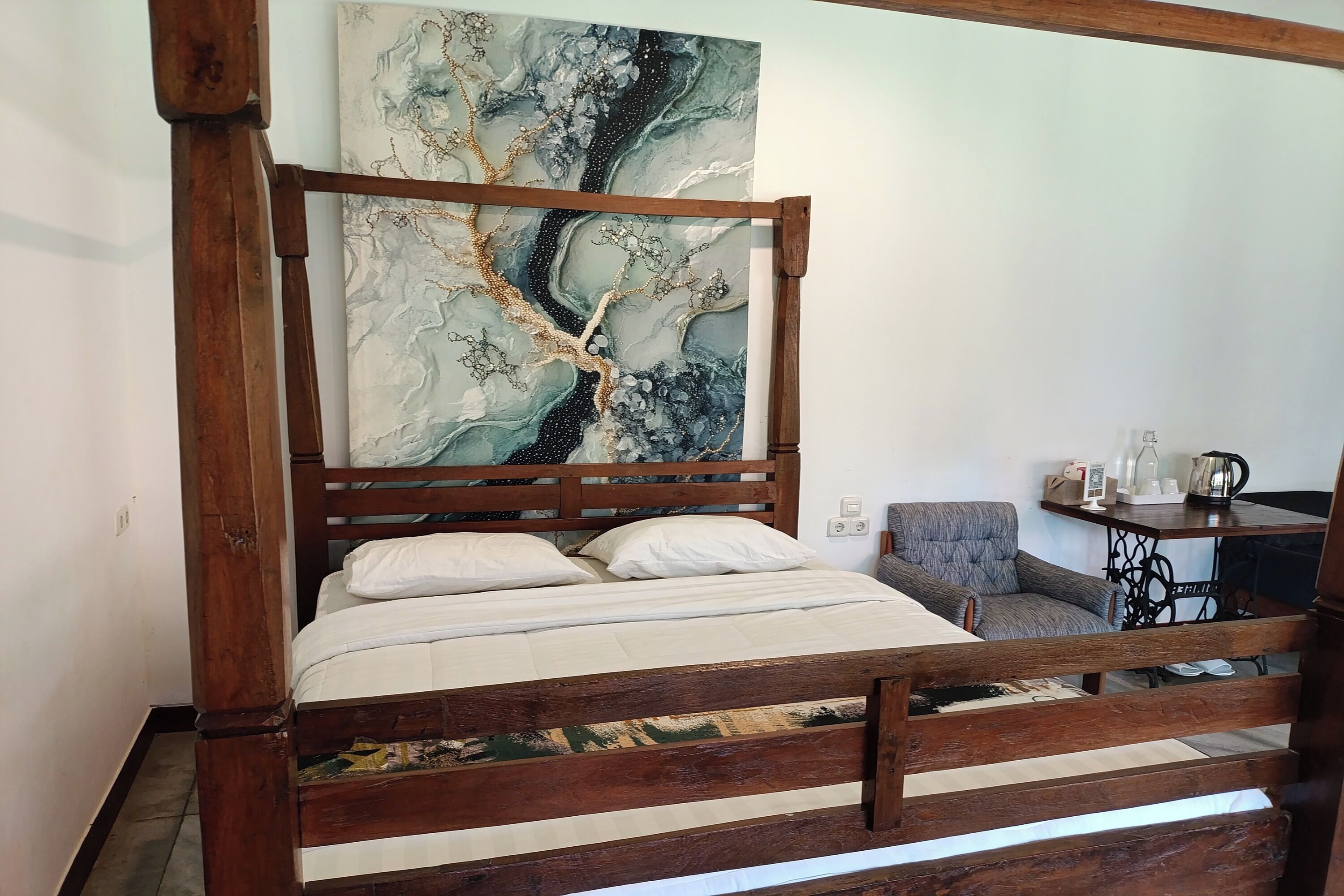 A wooden bed with a headboard, a painting above it, and a small table with items on it.