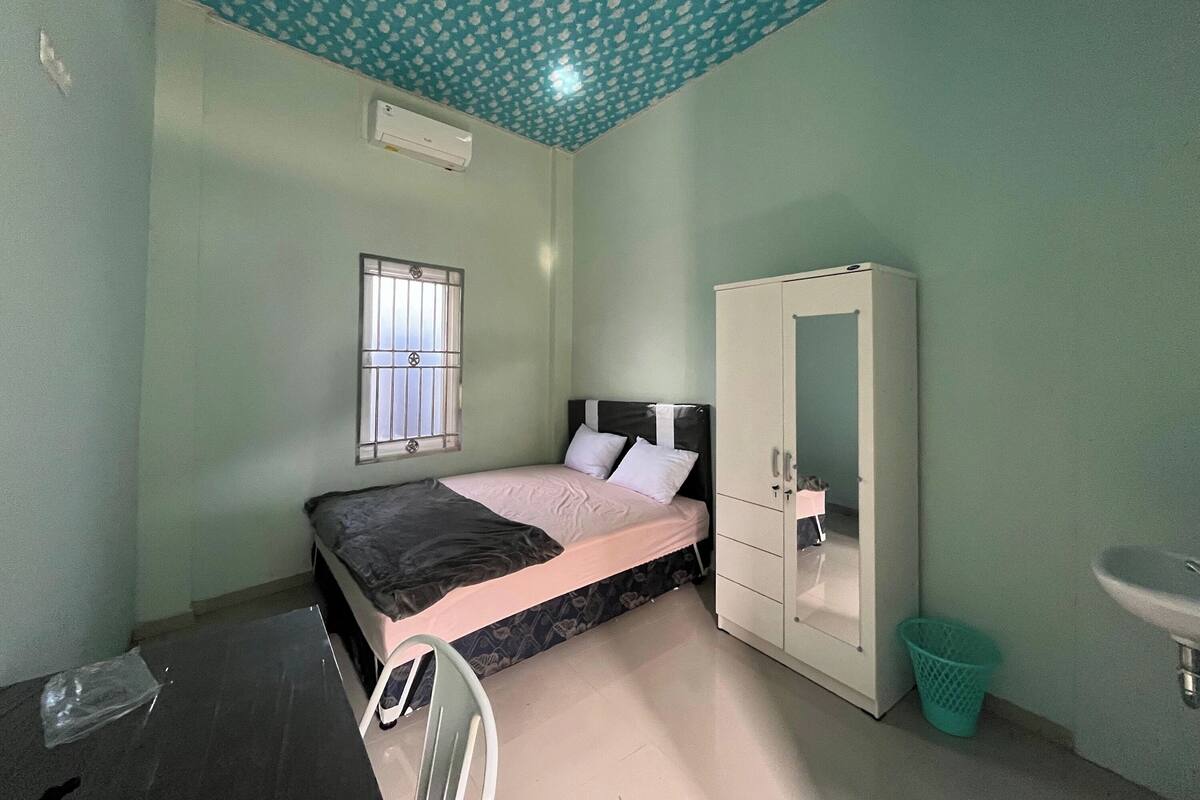 A bedroom with a bed, a wardrobe, a window with bars, a sink, and a trash bin.