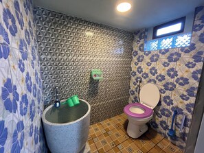 A bathroom with a geometric-patterned wall, a toilet with a purple seat, a large blue bathtub, and a green cleaning bucket. - Al-Barokah Embun Syariah (Palembang)