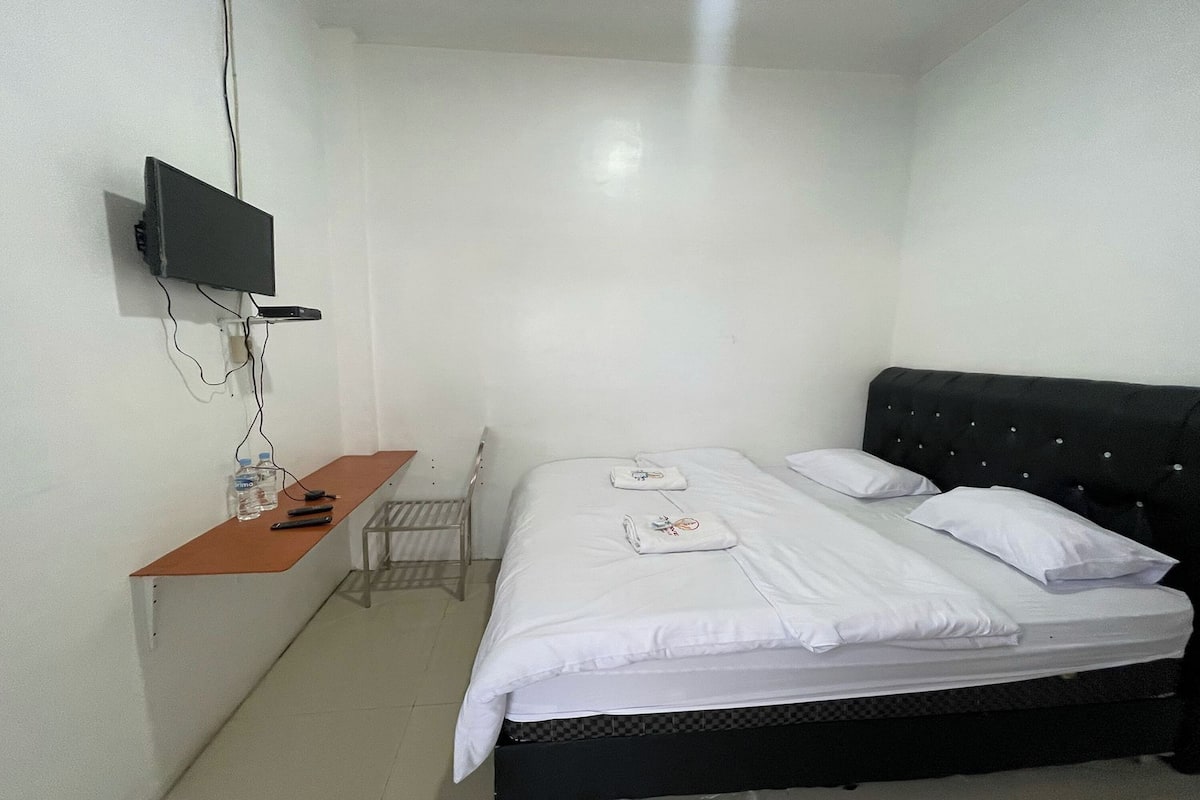 A bedroom with a bed, a TV mounted on the wall, a small table with a bottle and remote, and a chair.