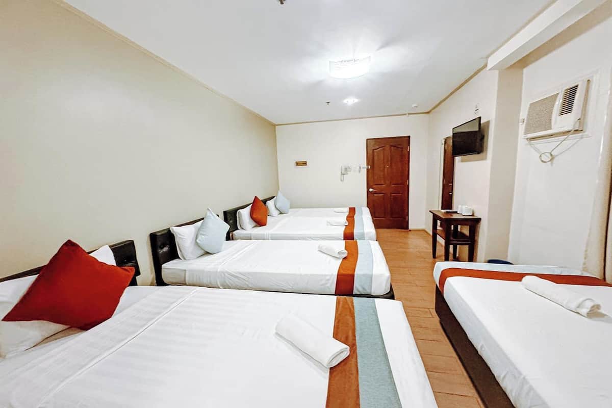A hotel room with four single beds, each with a pillow and a blanket. There is a wooden door, a small table with a chair, and a TV mounted on the wall.