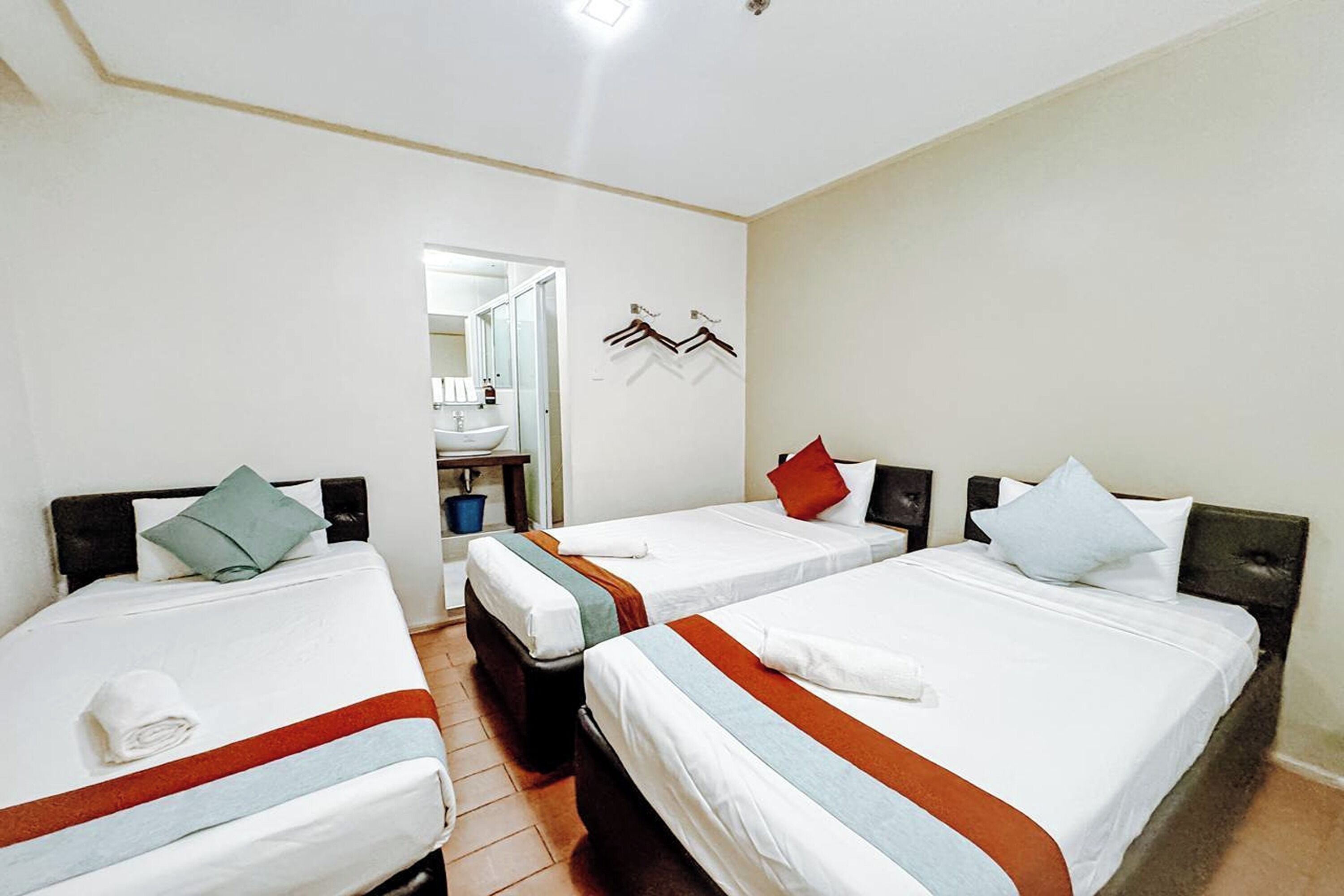 Une chambre d’hôtel avec trois lits simples, chacun étant équipé d’un oreiller et d’une couverture. On y trouve également une petite table avec une chaise et un miroir dans un coin.