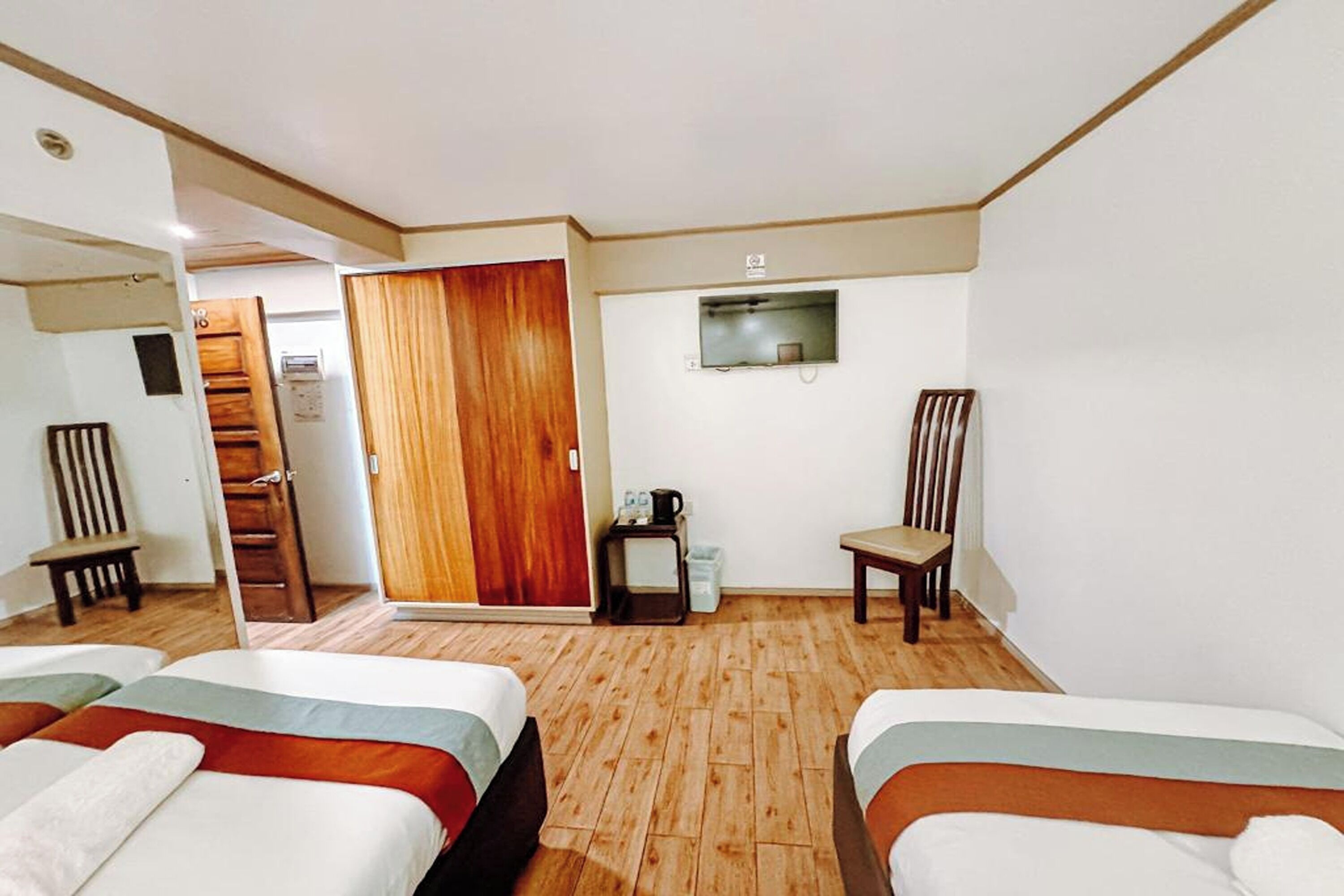 A hotel room with two beds, a wooden floor, a small table with a kettle, a chair, and a wooden door.