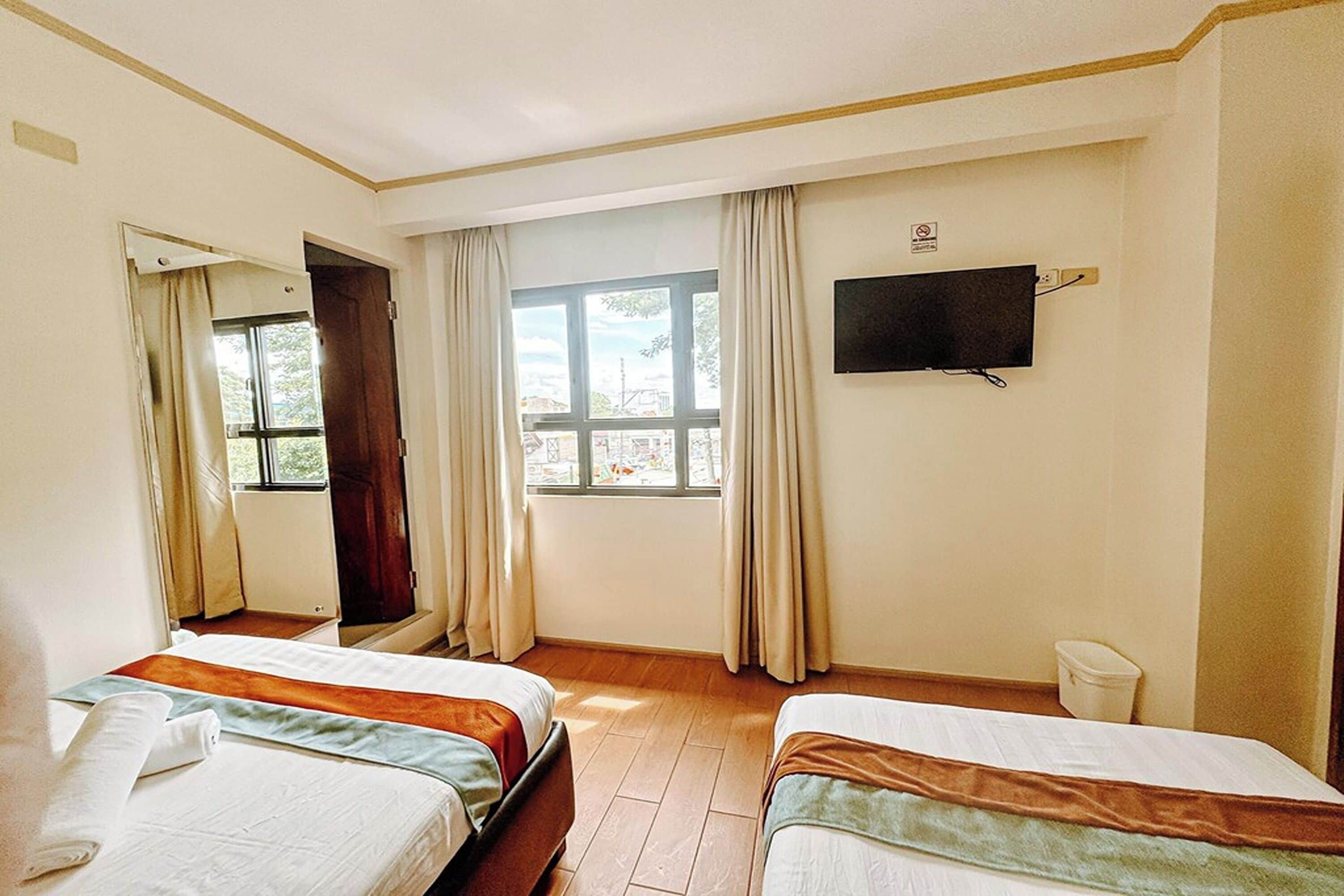 A hotel room with two beds, a window with curtains, a television on the wall, and a small trash bin.
