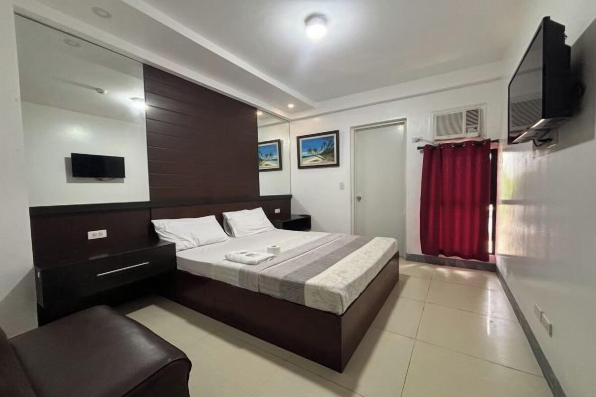 A bedroom with a large bed, a bedside table, a TV mounted on the wall, and a window with a red curtain.