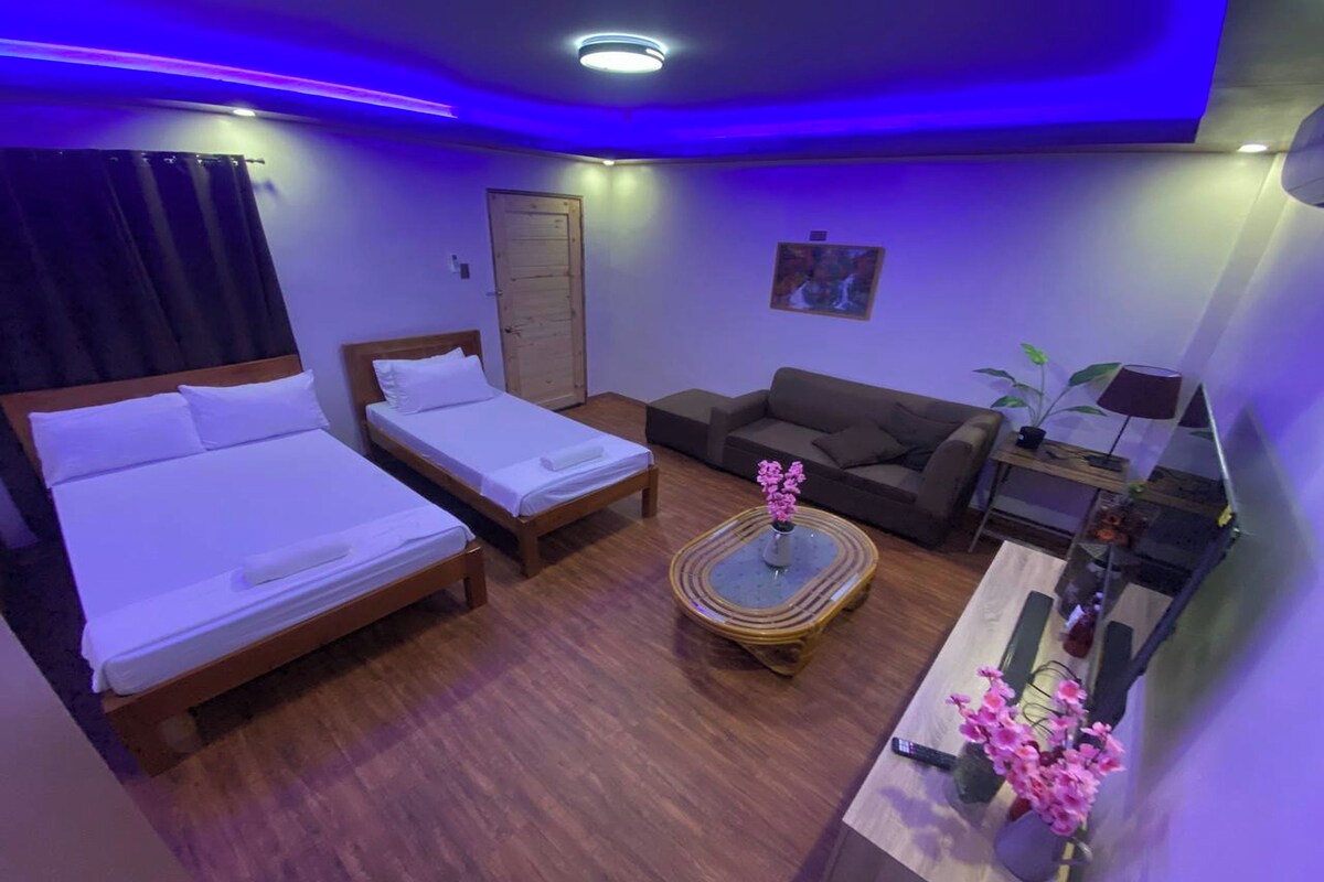 A hotel room with two single beds, a sofa, a small table with a vase of flowers, and a lamp.