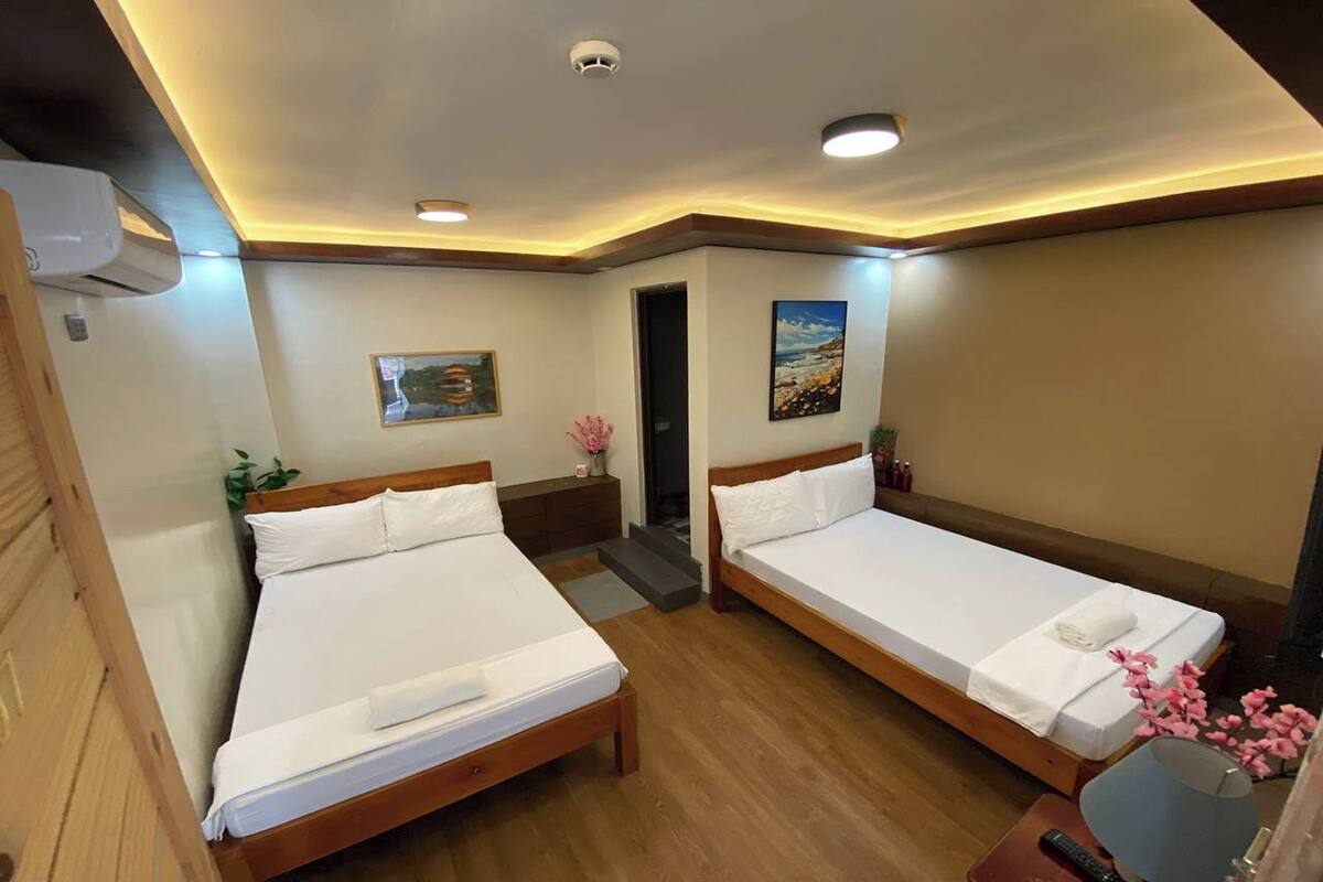 A hotel room with two single beds, wooden flooring, a ceiling-mounted air conditioner, a small desk with a lamp, and a painting on the wall.