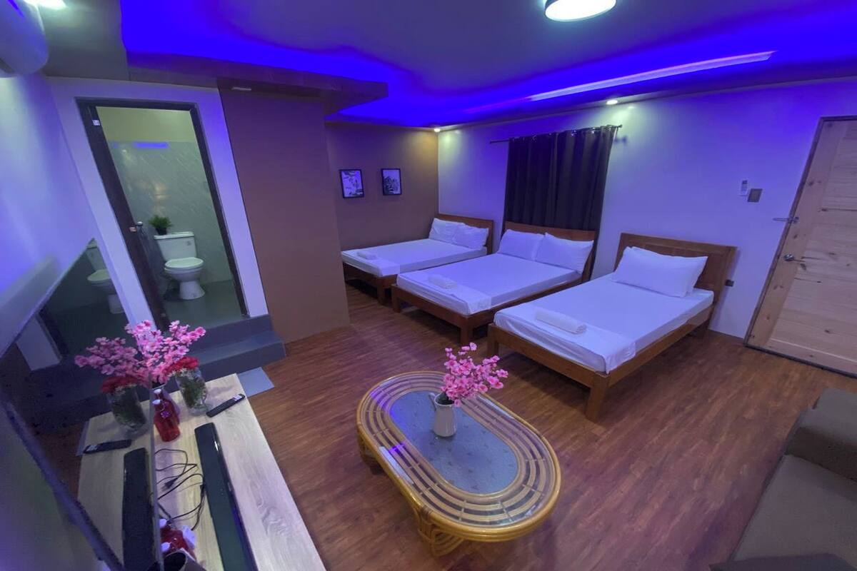 A hotel room with two single beds, a wooden floor, a small table with a vase of flowers, and a bathroom visible through a glass door.