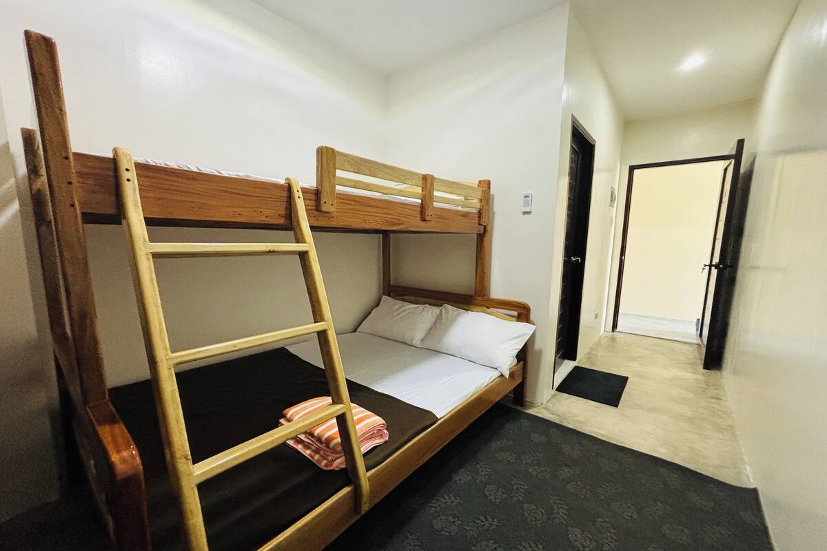 A bunk bed with a wooden ladder, a single bed with white bedding, a black mat at the foot of the bed, and a door in the background.