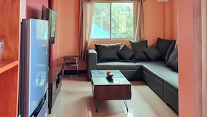 Un espacio habitable compacto con sofá, una mesita, refrigerador y una ventana con cortinas.