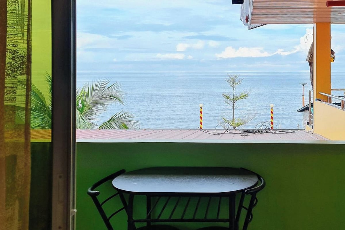 Blick vom Balkon mit Tisch und Stühlen auf ein ruhiges Meer und Palmen.