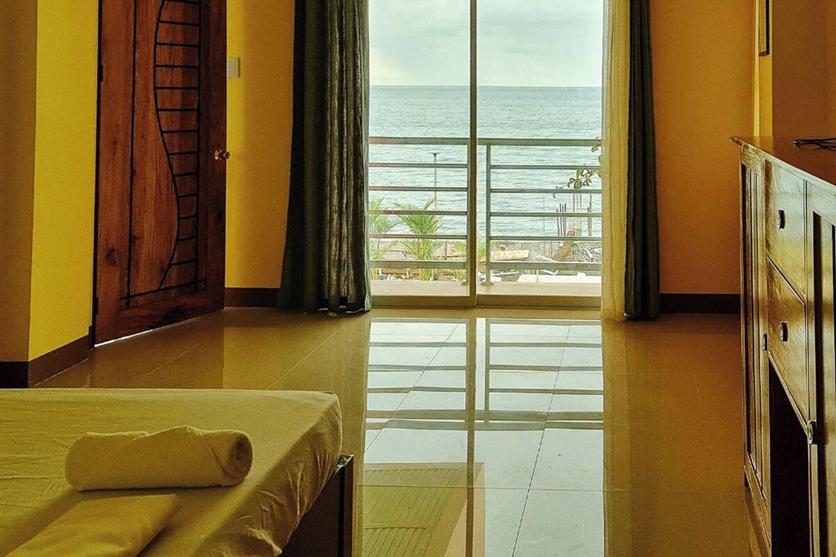 Quarto de hotel com uma cama, guarda-roupa de madeira, varanda com vista para o mar e porta com puxador de metal.