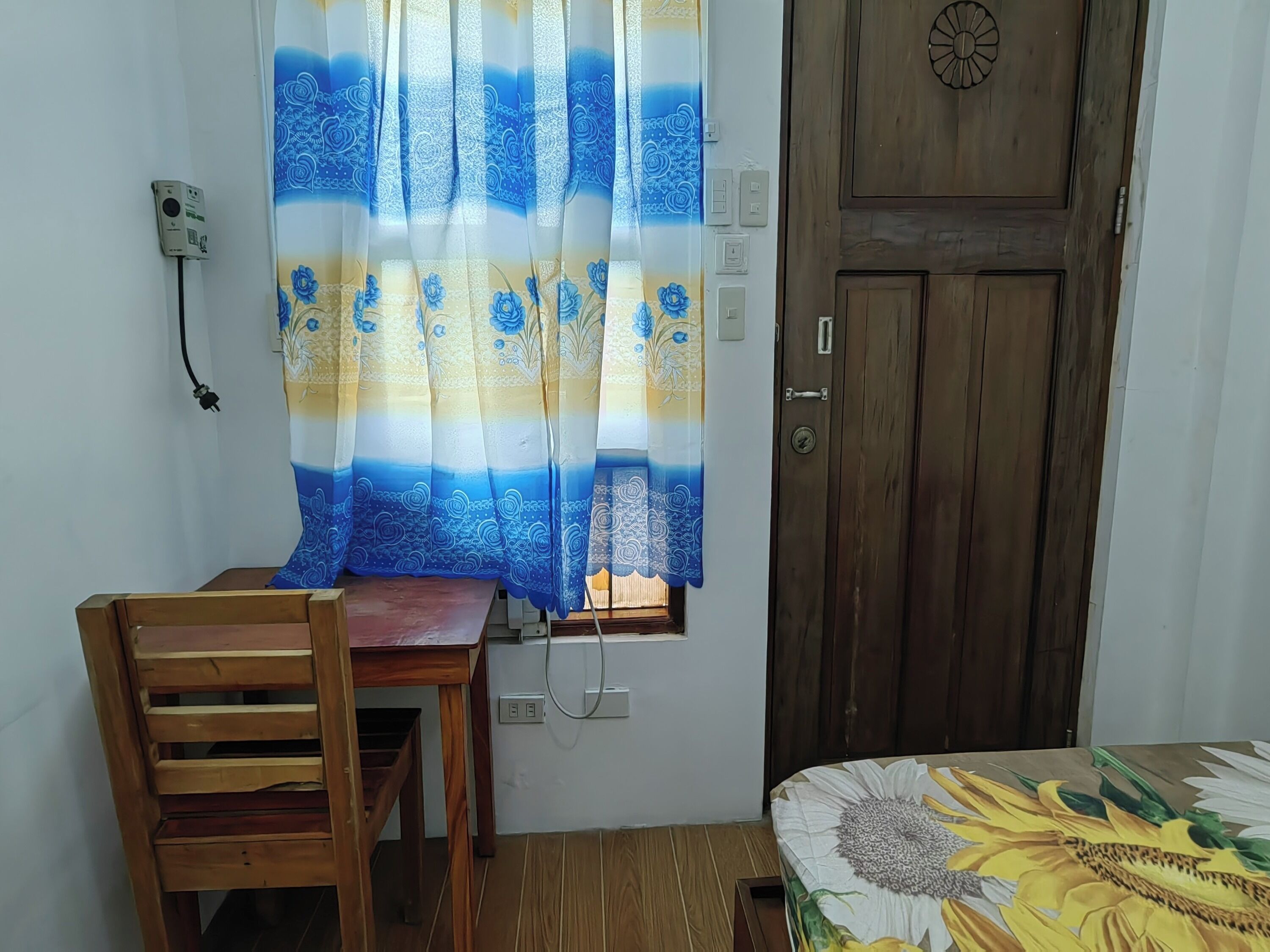A room with a wooden desk, a chair, a bed with a sunflower bedspread, a wooden door, and a window with a blue and white patterned curtain.