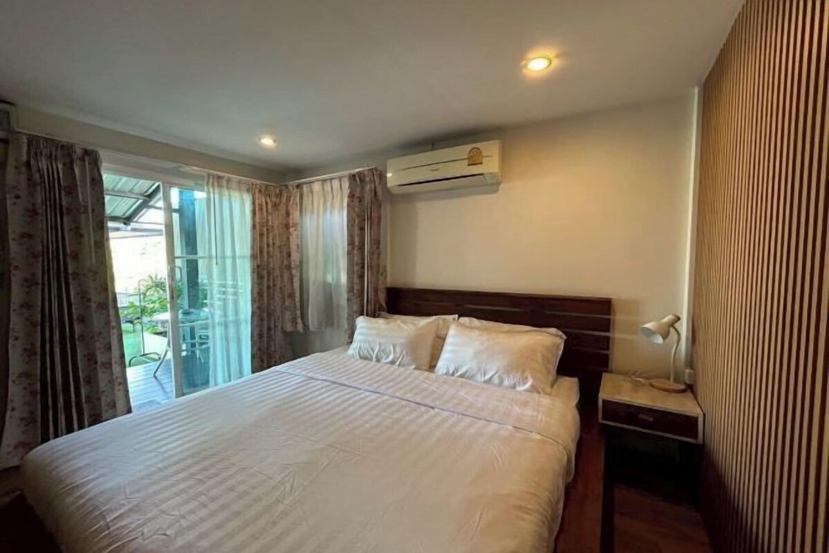 Habitación de hotel con una cama grande, un equipo de aire acondicionado, una mesita de noche con lámpara y un ventanal corredero que da a un área exterior.