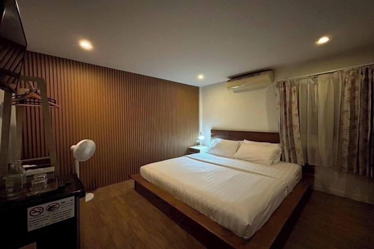 Habitación de hotel con pared de paneles de madera, cama con ropa blanca, mesita de noche con lámpara y ventana con cortinas.