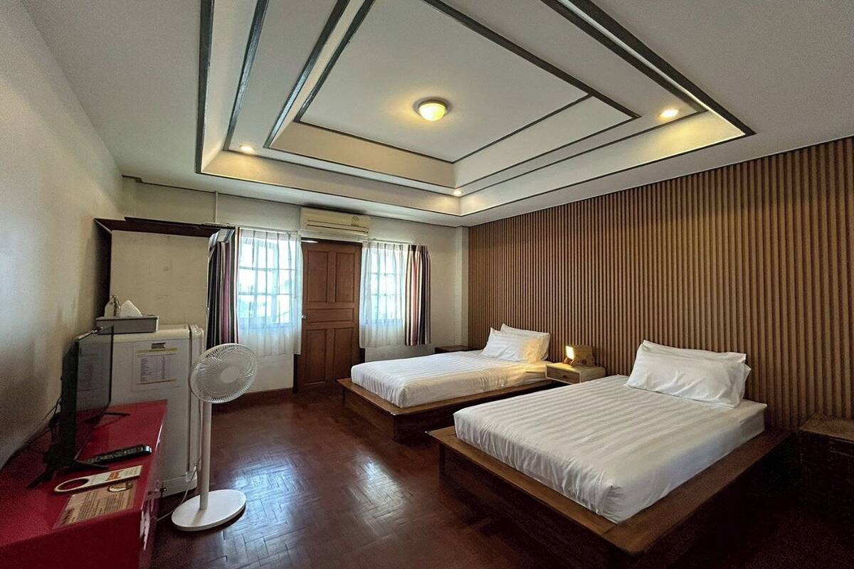 A hotel room with two single beds, a desk with a computer, a standing fan, and a radiator.