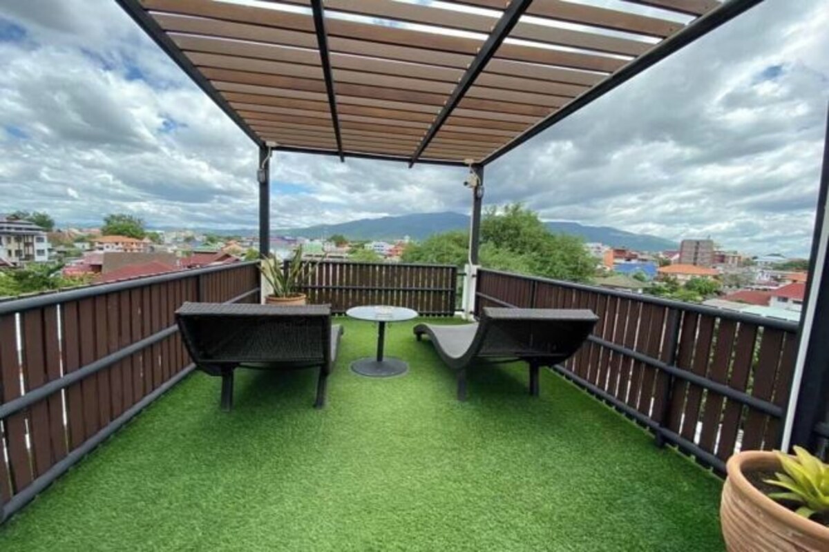 Un balcon avec un revêtement de gazon synthétique, deux fauteuils en osier noir, une petite table ronde et une pergola en bois.
