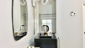 Habitación de hotel con encimera negra, un espejo, un ventilador blanco, un hervidor y una bandeja con una taza y un vaso.