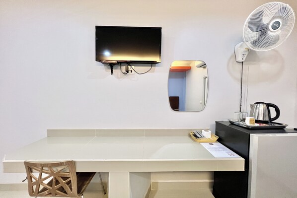 A hotel room with a flat-screen TV mounted on the wall, a standing fan, a small table with a kettle and a tray, and a chair. - Full House Udon (Udon Thani)