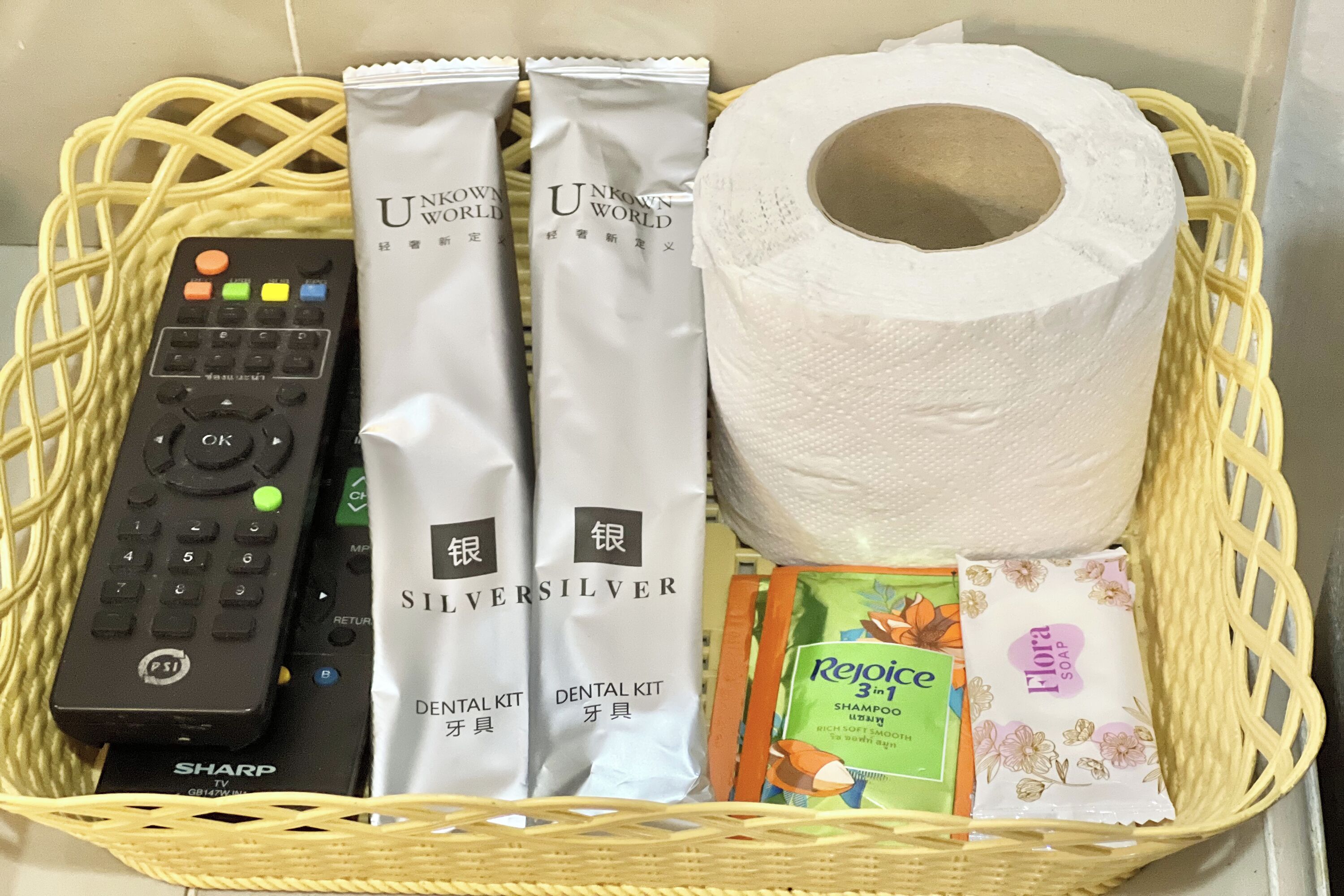 A basket containing a remote control, two dental kits, a roll of toilet paper, a shampoo bar, and a soap bar.