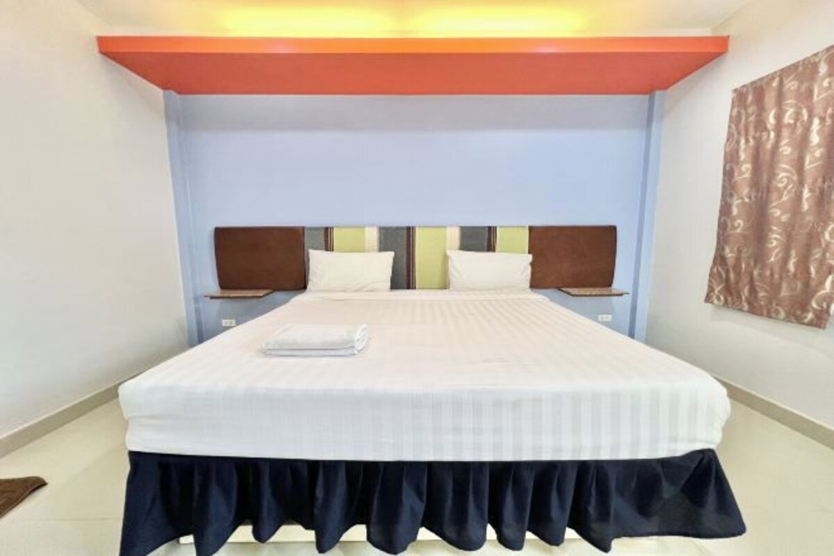 A neatly made bed with a white bedspread and a dark skirt at the foot. There are two bedside tables with lamps, and a wall-mounted shelf above the bed.