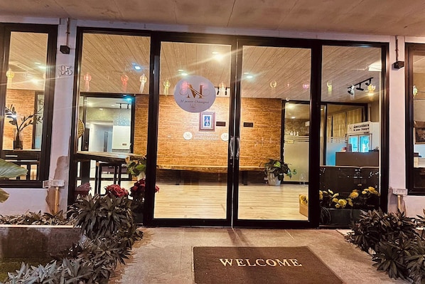 L’entrée d’un hôtel, avec un comptoir d’accueil, un paillasson où est écrit « Bienvenue » et des plantes d’intérieur.
