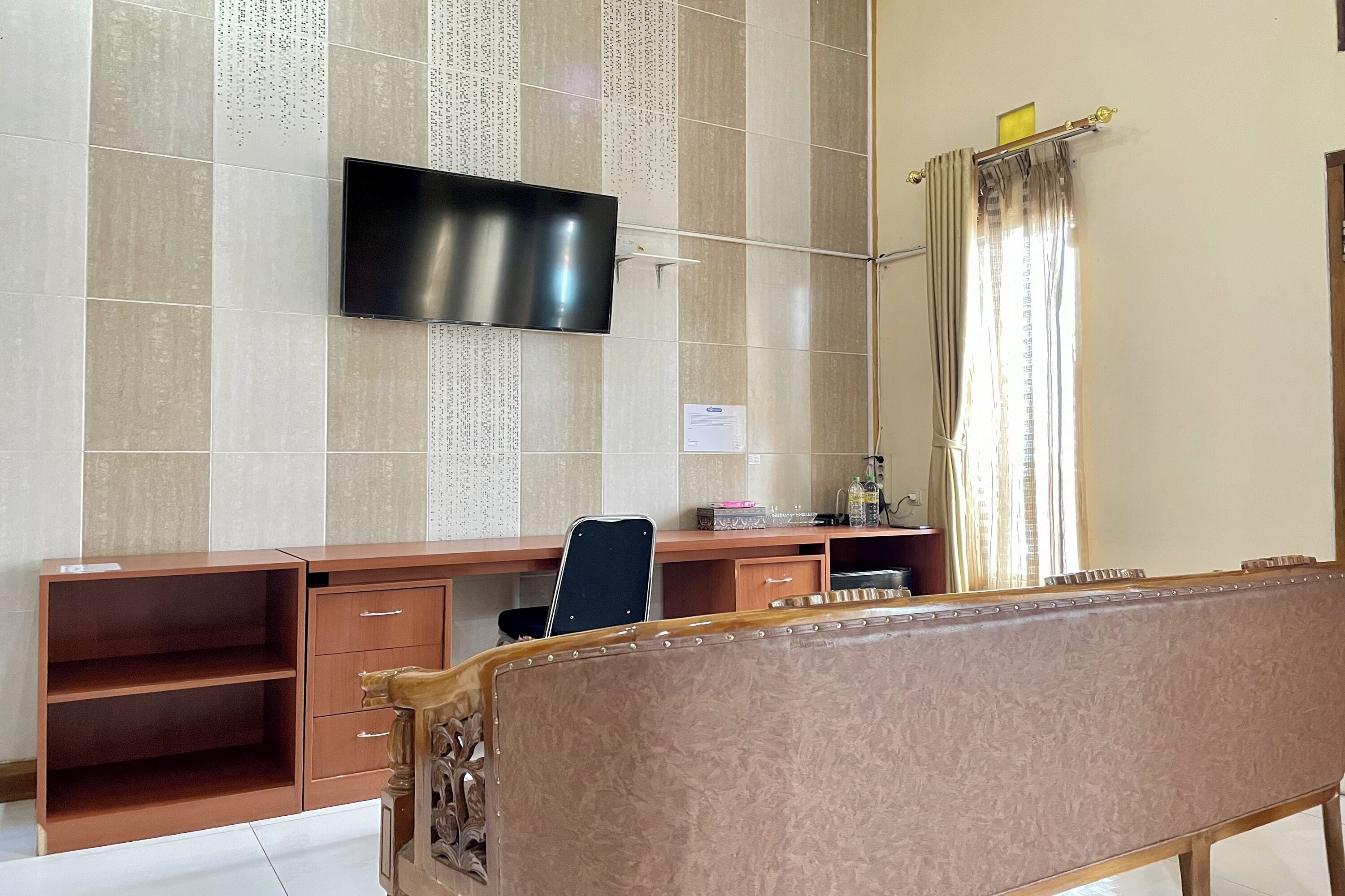 A room with a flat-screen TV mounted on the wall, a wooden desk with drawers, a chair, and a sofa.