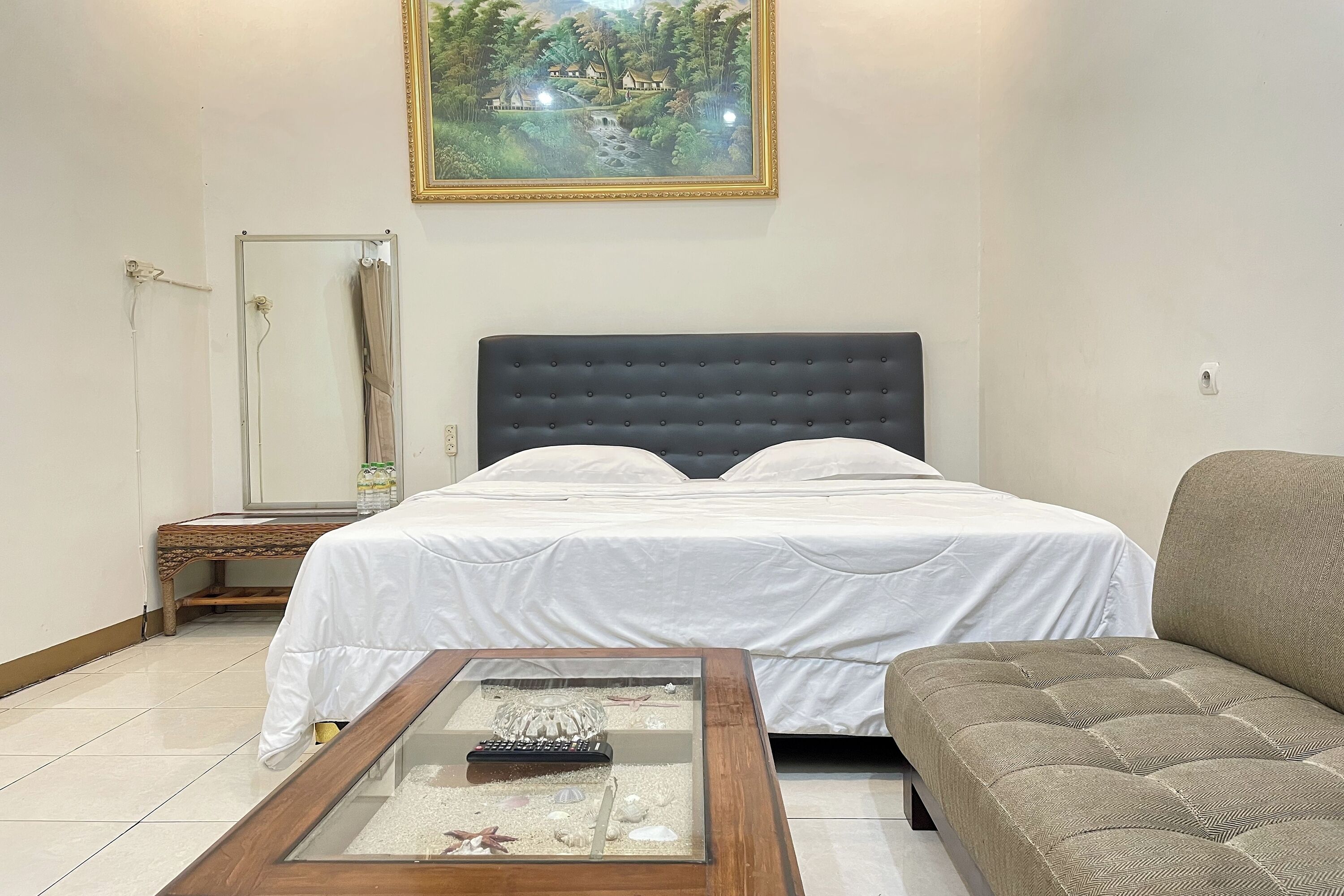A bedroom with a bed, a glass-top coffee table, a sofa, a painting on the wall, and a small table with a lamp.