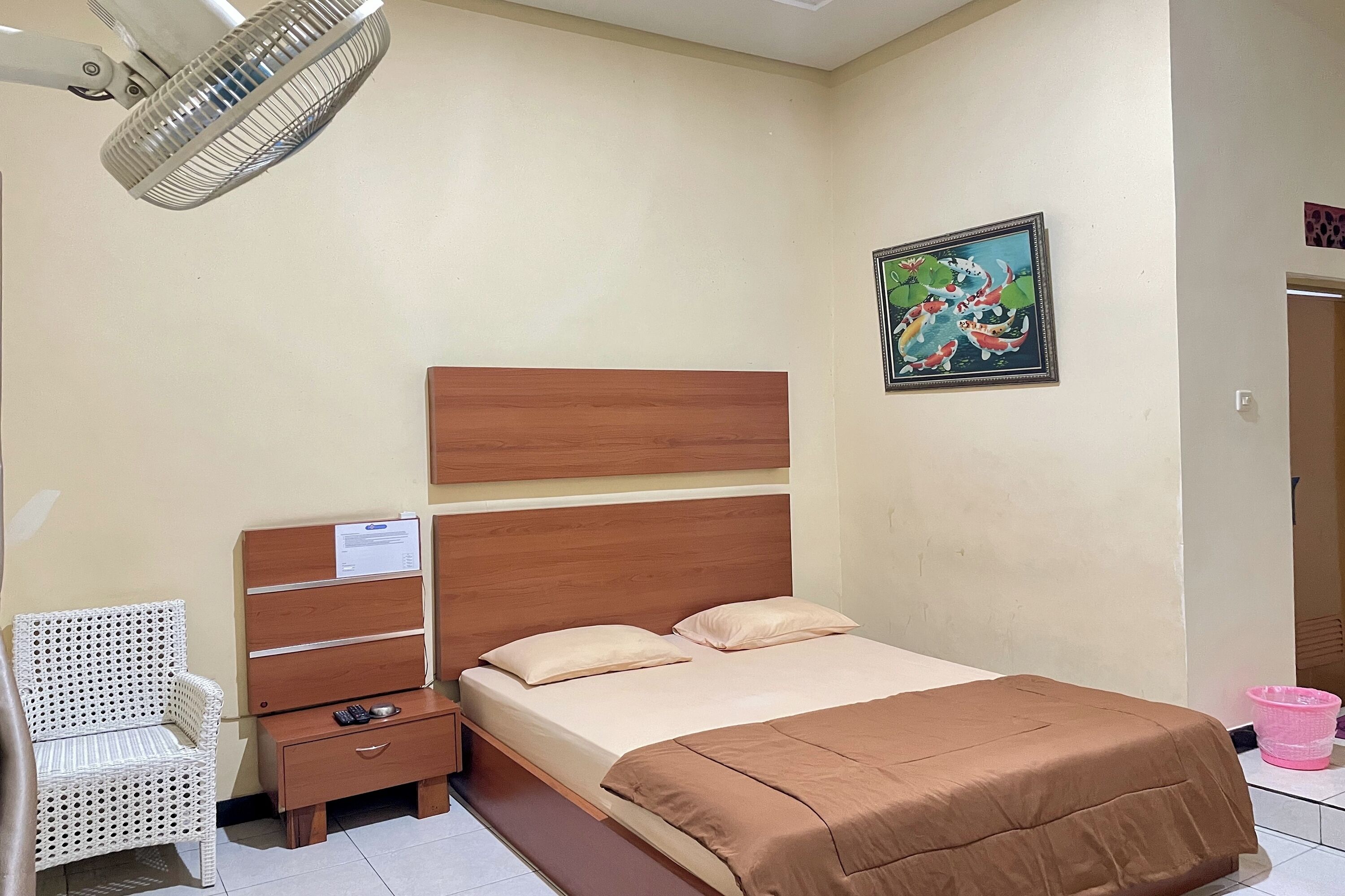 A bedroom with a bed, a nightstand, a chair, a wall-mounted fan, and a painting.